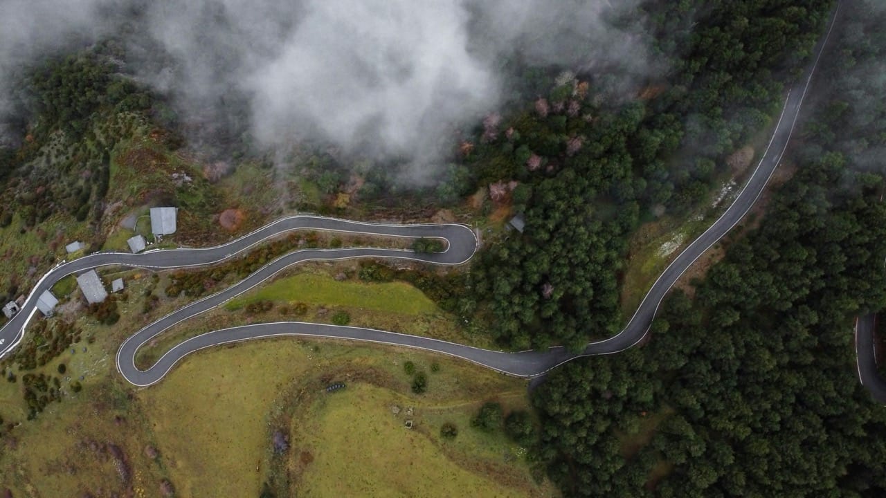 Vue aérienne d'une route sinueuse en Andorre, traversant un paysage montagneux verdoyant, partiellement enveloppé par des nuages bas, avec une ambiance sereine et mystérieuse.