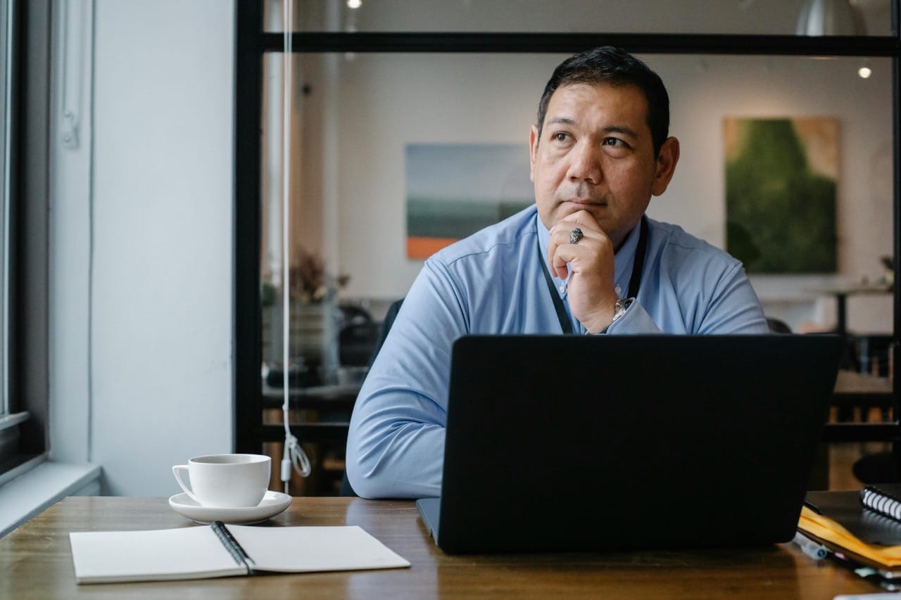 Homme en chemise bleue assis à son bureau, réfléchissant devant son ordinateur portable, avec une tasse de café et un carnet ouverts, dans un bureau moderne avec une ambiance professionnelle.