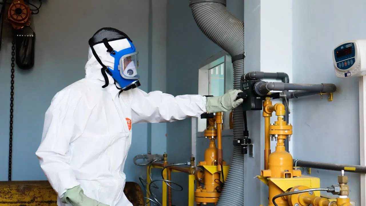 Technicien équipé d’une combinaison de protection intégrale et d’un masque filtrant, manipulant un système de tuyauterie dans un environnement industriel sensible.