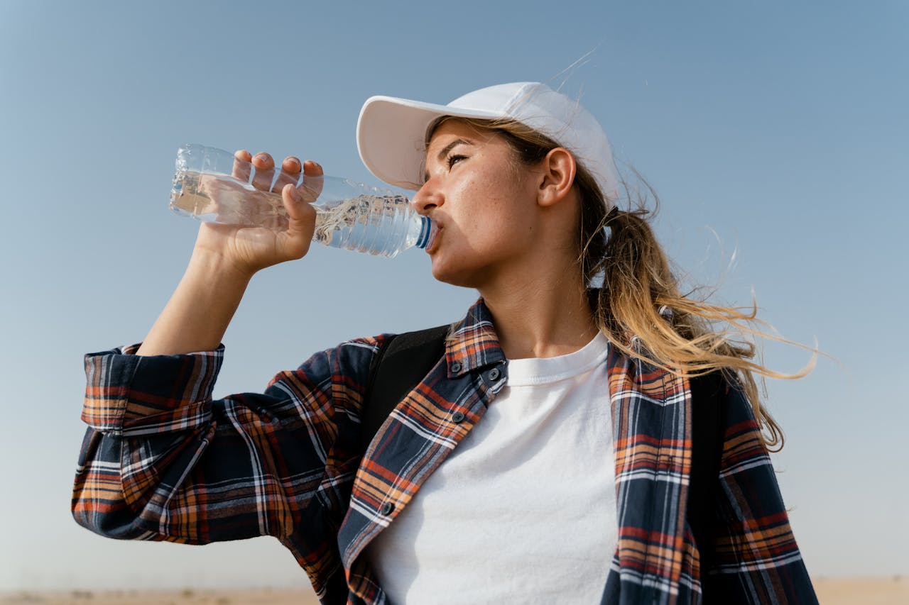 Jeune femme portant une casquette blanche et une chemise à carreaux, buvant de l’eau en plein soleil pour rester hydratée, dans un environnement chaud et sec.