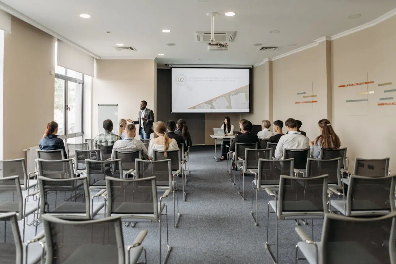 Conférence ou session de sensibilisation en entreprise, avec un intervenant debout devant un petit groupe de participants, dans une salle équipée d’un vidéoprojecteur et d’un écran de présentation.